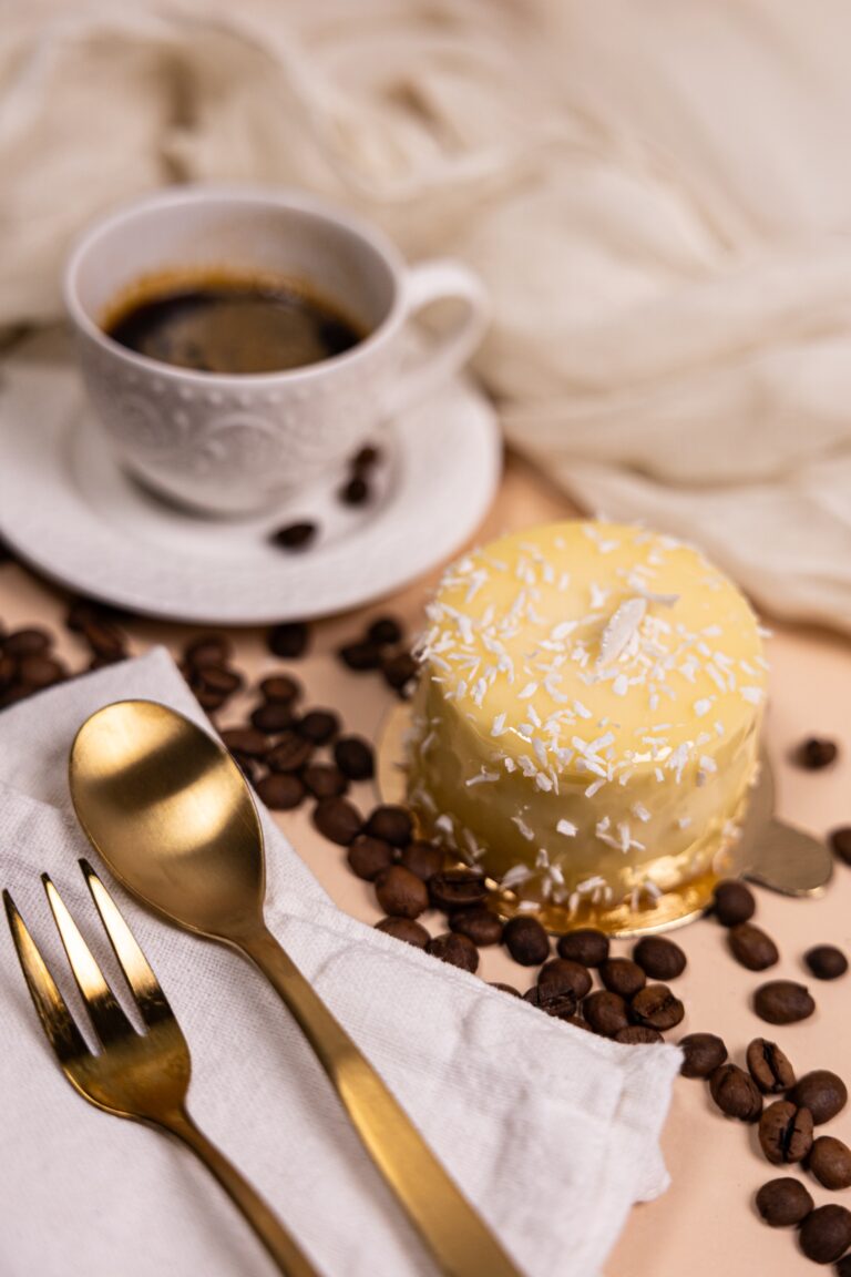 White-glazed cake with coconut flakes next to a cup of coffee and gold cutlery